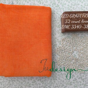 May include: A folded orange linen fabric with the text "Ju design" embroidered in the corner. A small wooden box with the text "RED GRAPEFRUIT 32 count linen DMC 3340-3883" is next to a cream-colored lace doily.