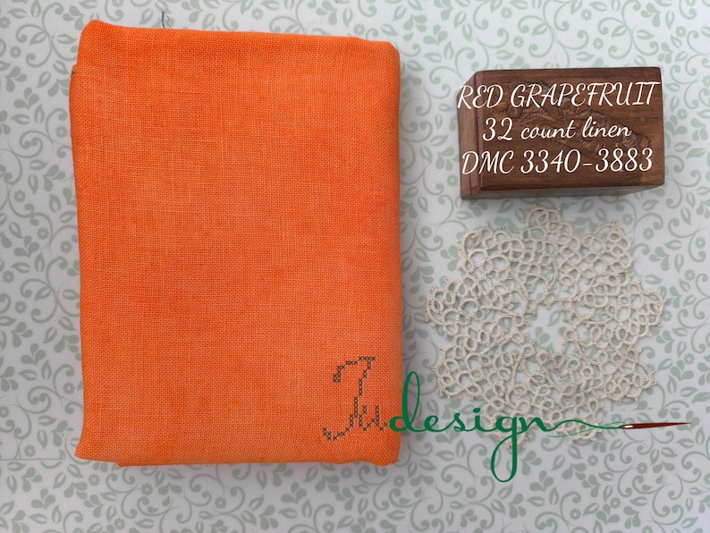 May include: A folded orange linen fabric with the text "Ju design" embroidered in the corner. A small wooden box with the text "RED GRAPEFRUIT 32 count linen DMC 3340-3883" is next to a cream-colored lace doily.