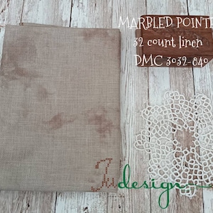 Op de afbeelding: Een stuk beige linnen stof met de tekst "Marbled Pointer 32 count linen DMC 3032-840" gedrukt op een houten blok.