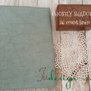 Puede incluir: Tela de lino verde salvia con diseño de punto de cruz y la palabra "Ju design". Una caja de madera con el texto "Ghostly Shadows 36 count linen" y un tapete de encaje blanco también están presentes.