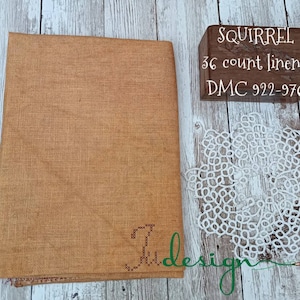 May include: A rectangular piece of tan linen fabric with a visible weave, featuring a small cross-stitch monogram in the corner. Next to it is a white lace doily and a wooden block with the text SQUIRREL 36 count linen DMC 922-976.