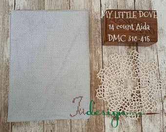 Peut inclure: Un tissu bleu clair pour point de croix avec une lettre "J" brodée. Un bloc de bois avec le texte "MY LITTLE DOVE 14 count Aida DMC 318-415" est à côté d'un napperon en dentelle blanche. Sur une surface en bois.