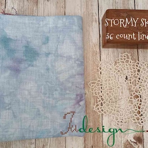 May include: A folded piece of light blue fabric with a watercolor effect, next to a wooden box labeled "Stormy Sky 36 count linen" and a white lace doily. The fabric has a purple and teal design.