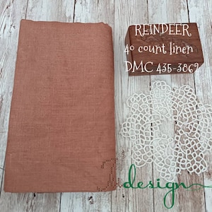 Puede incluir: Un trozo doblado de tela de lino rosa polvoriento junto a un bloque de madera con la palabra "REINDEER" y otro texto. Un tapete de encaje blanco y la palabra "design" en escritura verde también están en la imagen.