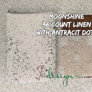 May include: A white linen fabric with dark gray spots, a wooden sign that reads "MOONSHINE 36 COUNT LINEN WITH ANTRACIT DOTS", and a cream-colored lace doily. The fabric has a stitched design.
