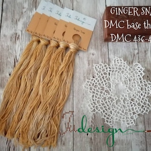 Puede incluir: Cinco carretes de hilo de bordar DMC de color marrón claro, etiquetados como "Ginger Snap" y "DMC 436-437". El hilo se utiliza para un encaje blanco, con el texto "design" visible en el fondo.