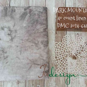 Op de afbeelding: Een stuk stof met een gemarmerd grijs en wit patroon, een houten doos met de tekst "DARK MOUNTAIN 40 count linen DMC 3024-648" en een witte kanten placemat. De stof heeft een kruissteekontwerp in de hoek.