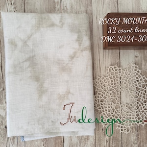 May include: A piece of off-white linen fabric with a brown embroidered letter "J" in the lower right corner. The fabric is labeled "Rocky Mountain 32 count linen DMC 3024-3032".