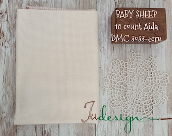 Peut inclure: Un tissu de point de croix crème avec une lettre "J" cousue dans le coin. Un bloc de bois affiche "BABY SHEEP 18 count Aida DMC 3033-ecru". Un napperon en dentelle blanche et le mot "design" sont également visibles.