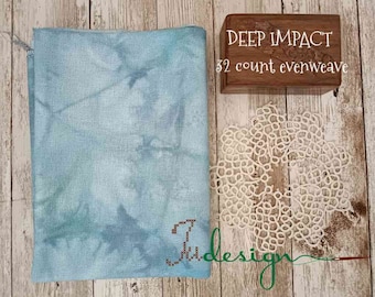 Puede incluir: Un trozo doblado de tela evenweave azul claro con efecto acuarela, junto a una caja de madera con el texto "DEEP IMPACT 32 count evenweave". También se ve un tapete de encaje blanco y la palabra "design".