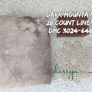 Op de afbeelding: Een stuk Dark Mountain 28 count linnen stof met een gevlekt grijs en beige patroon. Een houten blok met tekst en een crèmekleurig kanten kleedje staan ook in de afbeelding. De tekst op het blok luidt "DARK MOUNTAIN 28 COUNT LINEN DMC 3024-648".