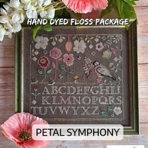 May include: A framed cross-stitch sampler on dark gray fabric featuring a bird, various flowers, and the alphabet in white thread. The text HAND DYED FLOSS PACKAGE and PETAL SYMPHONY are visible. The piece is surrounded by pink and white artificial flowers.