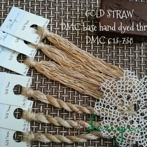Puede incluir: Hilo de bordar teñido a mano de color beige claro, etiquetado como "Gold Straw" y "DMC 613-738". El hilo está enrollado en madejas y se muestra en tarjetas blancas. También se muestra una pequeña caja de madera con la etiqueta "GOLD STRAW DMC base hand dyed thread". Un encaje blanco está en primer plano.