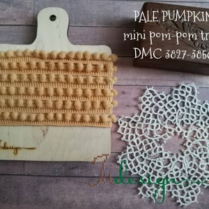 May include: Pale pumpkin mini pom-pom trim displayed on a wooden cutting board. The trim is a light orange color and is wrapped around the board. A wooden box with text and a white lace doily are also in the image.