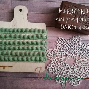 Può includere: Un tagliere in legno mostra un rotolo di mini passamaneria con pompon verde menta. La passamaneria è disposta in file ordinate. Sono presenti anche una piccola scatola di legno con la scritta "MERRY GREEN" e un centrino di pizzo bianco.