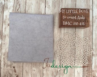 Peut inclure: Un tissu gris clair pour point de croix, une boîte en bois avec le texte "MY LITTLE DOVE 20 count Aida DMC 318-415", et un napperon en dentelle crème sont disposés sur une surface en bois. Le tissu est plié proprement, et le napperon a un design ajouré délicat.