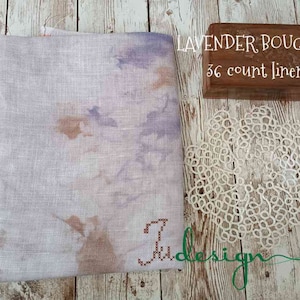 May include: A folded piece of lavender-colored linen fabric with brown and purple accents. The text "LAVENDER BOUQUET 36 count linen" is visible. A small wooden box and a white lace doily are also present.