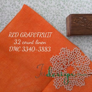 May include: An orange linen fabric square with the text "RED GRAPEFRUIT 32 count linen DMC 3340-3883". A small, carved wooden box and a white lace doily are also present. The background has a floral pattern.