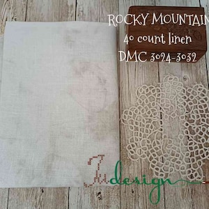 Op de afbeelding: Een witte linnen stof met een bruine houten doos waarop "Rocky Mountain 40 count linen DMC 3024-3032" staat en een witte kantonderlegger.