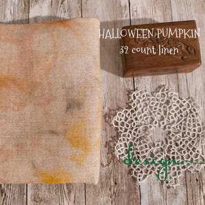 May include: A folded piece of linen fabric with orange and yellow watercolor-like patterns. A small wooden box with the text "HALLOWEEN PUMPKIN 32 count linen" is next to a white lace doily. The items are on a wooden surface.