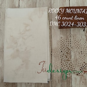 May include: A folded off-white linen fabric with a subtle brown pattern, a small embroidered letter, and a decorative doily. The text "ROCKY MOUNTAIN 46 count linen DMC 3024-3032" is visible on a wooden block.