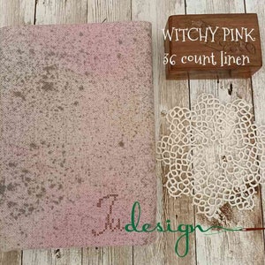 Puede incluir: Un trozo de tela de lino rosa con un diseño moteado, una caja de madera con el texto "Witchy Pink 36 count linen" y un tapete de encaje blanco. La tela tiene la palabra "Judesign" cosida en verde.