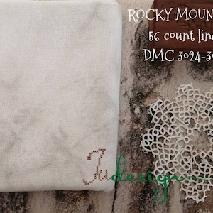 Op de afbeelding: Een stuk wit linnen stof met de tekst "ROCKY MOUNTAIN 56 count linen DMC 3024-3032" gedrukt op een bruin houten blok. Een wit kantonderlegger ligt op de tafel naast de stof.