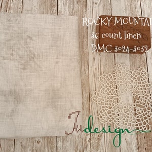 Op de afbeelding: Een gevouwen, gebroken witte linnen stof met een kruissteekontwerp in de hoek. Een witte, cirkelvormige kanten doily ligt naast de stof. Tekst op de afbeelding luidt "Rocky Mountain 36 count linen DMC 3024-3032".