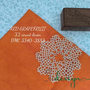 May include: An orange linen fabric square with the text "RED GRAPEFRUIT 32 count linen DMC 3340-3883" and a white lace doily. A small wooden box with carved details sits nearby.