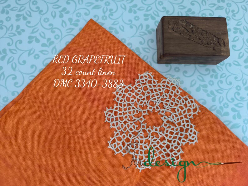 May include: An orange linen fabric square with the text "RED GRAPEFRUIT 32 count linen DMC 3340-3883" and a white lace doily. A small wooden box with carved details sits nearby.