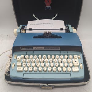 May include: A blue Smith-Corona Coronet Automatic 10 electric typewriter with a QWERTY keyboard, cream-colored keys, and a white paper sheet inserted in the carriage. The typewriter is housed in a black carrying case with a red emblem on the lid.