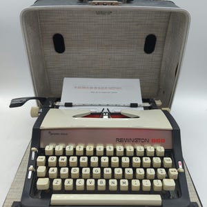 May include: Vintage Remington 666 typewriter in its original case. The typewriter has a cream-colored body with black keys and a black frame. The case is gray with a black handle. A sheet of paper is in the typewriter.