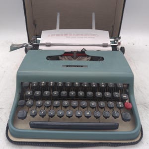 May include: A vintage teal typewriter with black keys and a red return key. The typewriter is open, revealing a sheet of paper with text. The model name "Lettera 22" is visible on the front. The typewriter is in a black carrying case.