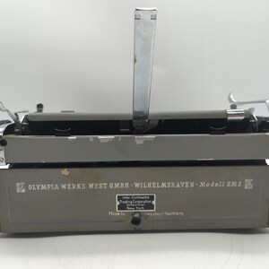 Pu&ograve; includere: Una macchina da scrivere grigia con una leva di ritorno carrello argentata e una leva argentata sul lato sinistro. La macchina da scrivere ha il testo "OLYMPIA WERKE WEST GMBH WILHELMSHAVEN Modell SM2" stampato sul davanti.