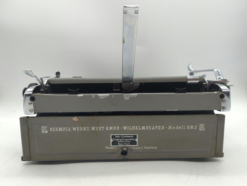 Pu&ograve; includere: Una macchina da scrivere grigia con una leva di ritorno carrello argentata e una leva argentata sul lato sinistro. La macchina da scrivere ha il testo "OLYMPIA WERKE WEST GMBH WILHELMSHAVEN Modell SM2" stampato sul davanti.