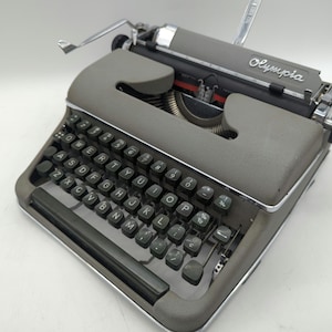 Pu&ograve; includere: Una macchina da scrivere vintage Olympia grigia con una tastiera nera e accenti argentati. La macchina da scrivere ha una leva di ritorno carrello e una leva di rilascio della carta.