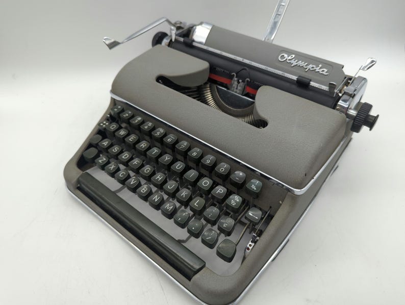 Pu&ograve; includere: Una macchina da scrivere vintage Olympia grigia con una tastiera nera e accenti argentati. La macchina da scrivere ha una leva di ritorno carrello e una leva di rilascio della carta.