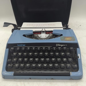 May include: A light blue Brother Charger 11 typewriter with black keys. The typewriter has a black carrying case. The keys are arranged in a QWERTY layout. A sheet of paper is inserted into the typewriter.