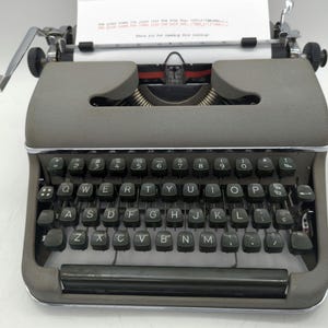 Pu&ograve; includere: Una macchina da scrivere vintage grigia con una tastiera nera e un foglio di carta bianco nel carrello. I tasti sono etichettati con le lettere QWERTYUIOPASDFGHJKLZXCVBNM e i numeri 2, 3, 4, 5, 6, 7, 8, 9, 0.
