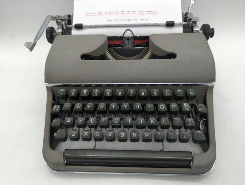 Pu&ograve; includere: Una macchina da scrivere vintage grigia con una tastiera nera e un foglio di carta bianco nel carrello. I tasti sono etichettati con le lettere QWERTYUIOPASDFGHJKLZXCVBNM e i numeri 2, 3, 4, 5, 6, 7, 8, 9, 0.