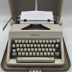 May include: A beige Olympia De Luxe portable typewriter with a QWERTY keyboard featuring cream-colored keys and green accent keys. A sheet of white paper is inserted into the carriage, displaying typed text in black and red ink.
