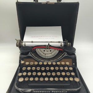 May include: A black vintage Underwood Portable typewriter in a black carrying case. The typewriter has a red ribbon and a sheet of paper with the text "The only time to start diet is the day you decide to."
