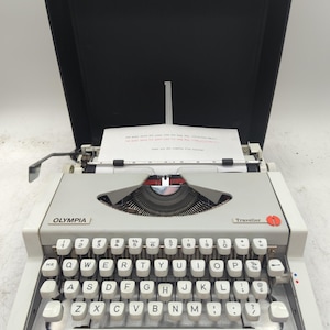 May include: An Olympia Traveller portable typewriter in a light gray casing with a blue front accent bar and a black carrying case lid. The QWERTY keyboard features white keys with black lettering, and a sheet of paper is loaded into the carriage.
