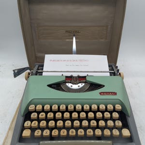 May include: A vintage Royal portable typewriter in a light green finish with cream-colored keys, housed in its original tan carrying case. A sheet of paper is inserted into the carriage, featuring typed text that reads, "The quick brown fox jumps over the lazy dog. 1234567890. How are you doing this morning?"