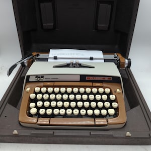 May include: A vintage Smith-Corona Classic 12 typewriter in its brown carrying case. The typewriter has a cream-colored body with brown accents and white keys. A sheet of paper is in the carriage. The keys are arranged in a QWERTY layout.
