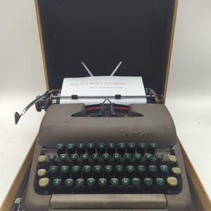 May include: A vintage Smith-Corona typewriter in its carrying case. The typewriter is brown with green and yellow keys. A sheet of paper is in the typewriter with text. The case is tan with a dark brown interior.