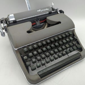 Pu&ograve; includere: Una macchina da scrivere vintage Olympia grigia con una tastiera nera e tasti verdi. La macchina da scrivere ha una leva di ritorno carrello argentata e una leva argentata sul lato sinistro.