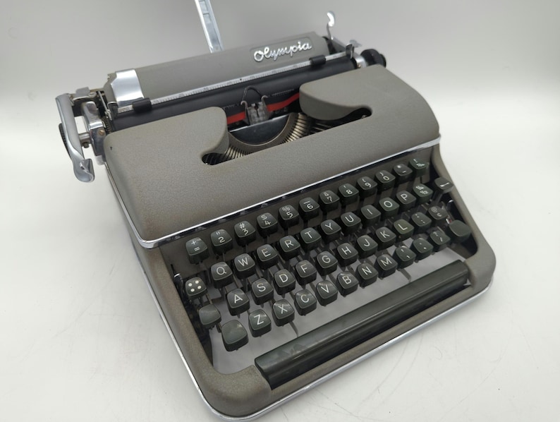 Pu&ograve; includere: Una macchina da scrivere vintage Olympia grigia con una tastiera nera e tasti verdi. La macchina da scrivere ha una leva di ritorno carrello argentata e una leva argentata sul lato sinistro.
