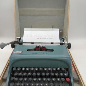 May include: A blue vintage Underwood Olivetti Lettera 32 typewriter in a beige carrying case. The typewriter has a standard QWERTY keyboard and a red return key. The case has a gold latch.