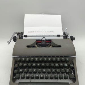 Pu&ograve; includere: Una macchina da scrivere vintage grigia con una tastiera nera e un foglio di carta nel carrello. Il testo sul foglio dice "The quick brown fox jumps over the lazy dog.  abcdefghijklmnopqrstuvwxyz!" e "Thank you for viewing this listing!"